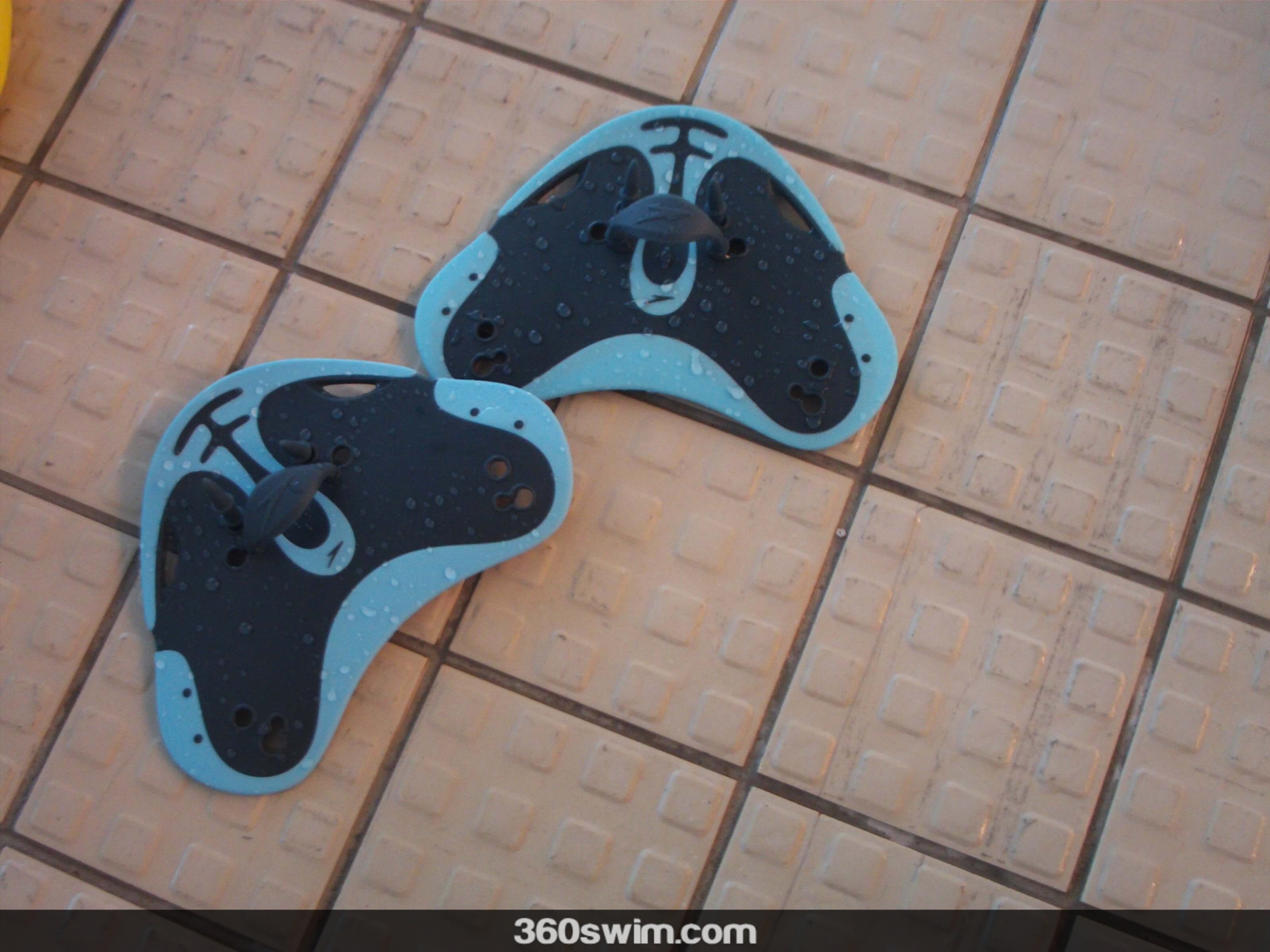 A pair of small, ergonomic, blue and black swimming paddles resting on the side of a pool.