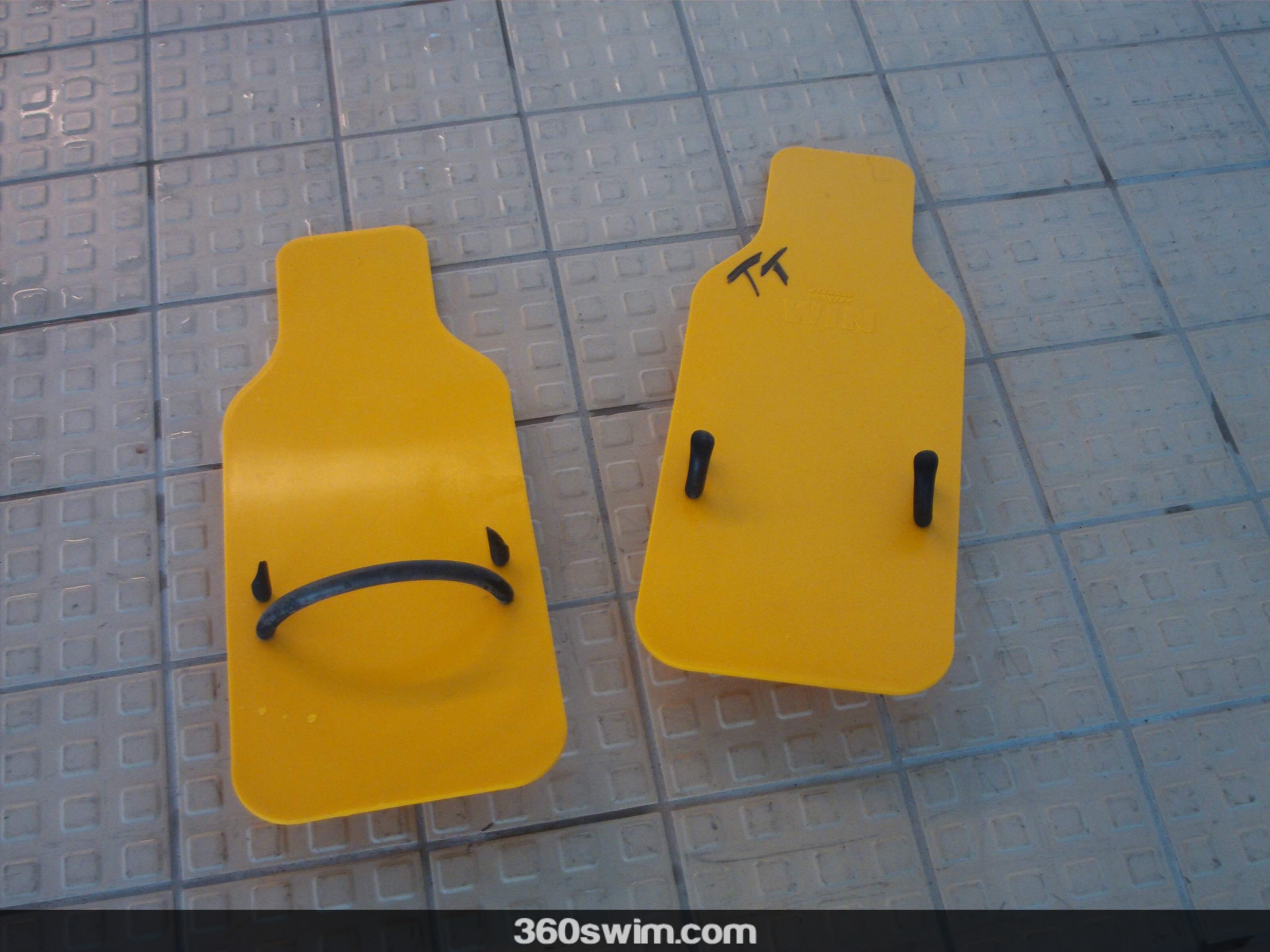 A pair of yellow, rectangular backstroke paddles lying on a tiled pool deck.